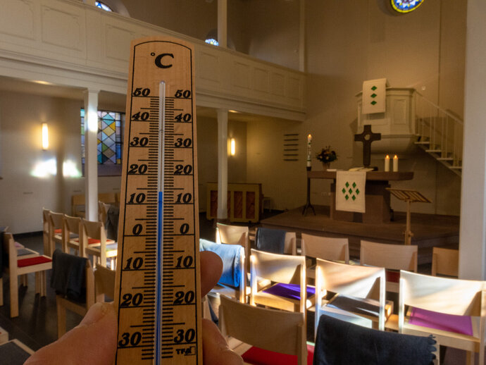 Energiesparen in der Kirche. In der Nazarethkirche in Frankfurt Eckenheim liegen während des Gottesdienstes Decken auf den Stühlen für die Gottesdienstbesucher bereit. Blick auf die Empore, Stuhlreihen mit den Decken. Seitlicher Lichteinfall durch die Fenster auf Fußboden und Stühle. Altar mit Kreuz und Kerzen - dahinter die Kanzel. Im Vordergrund hält eine Hand ein Thermometer mit Temperaturskala.